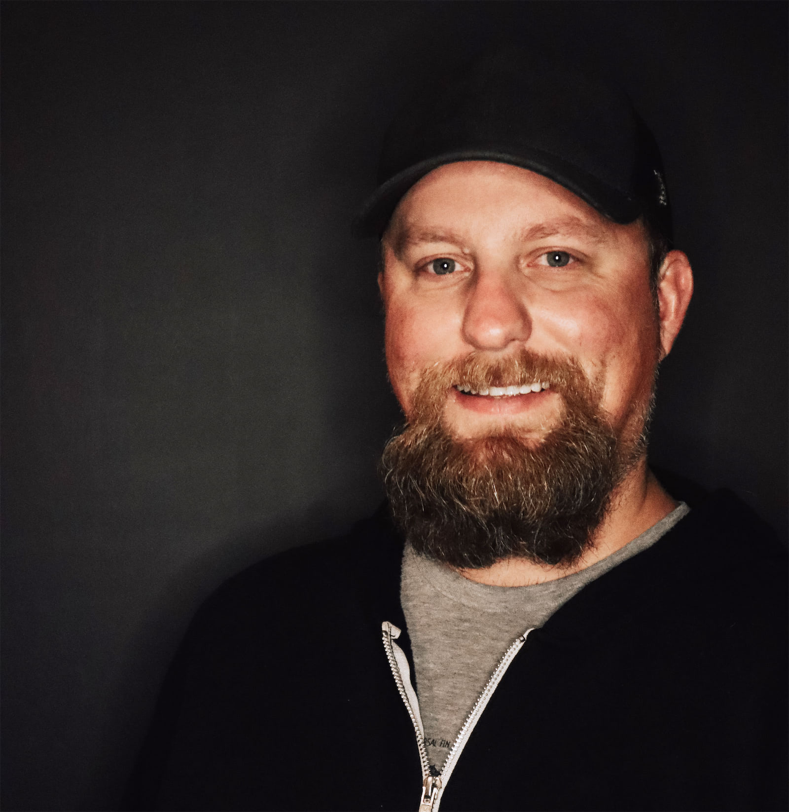 Smiling bearded man in a black cap and jacket against a dark background.